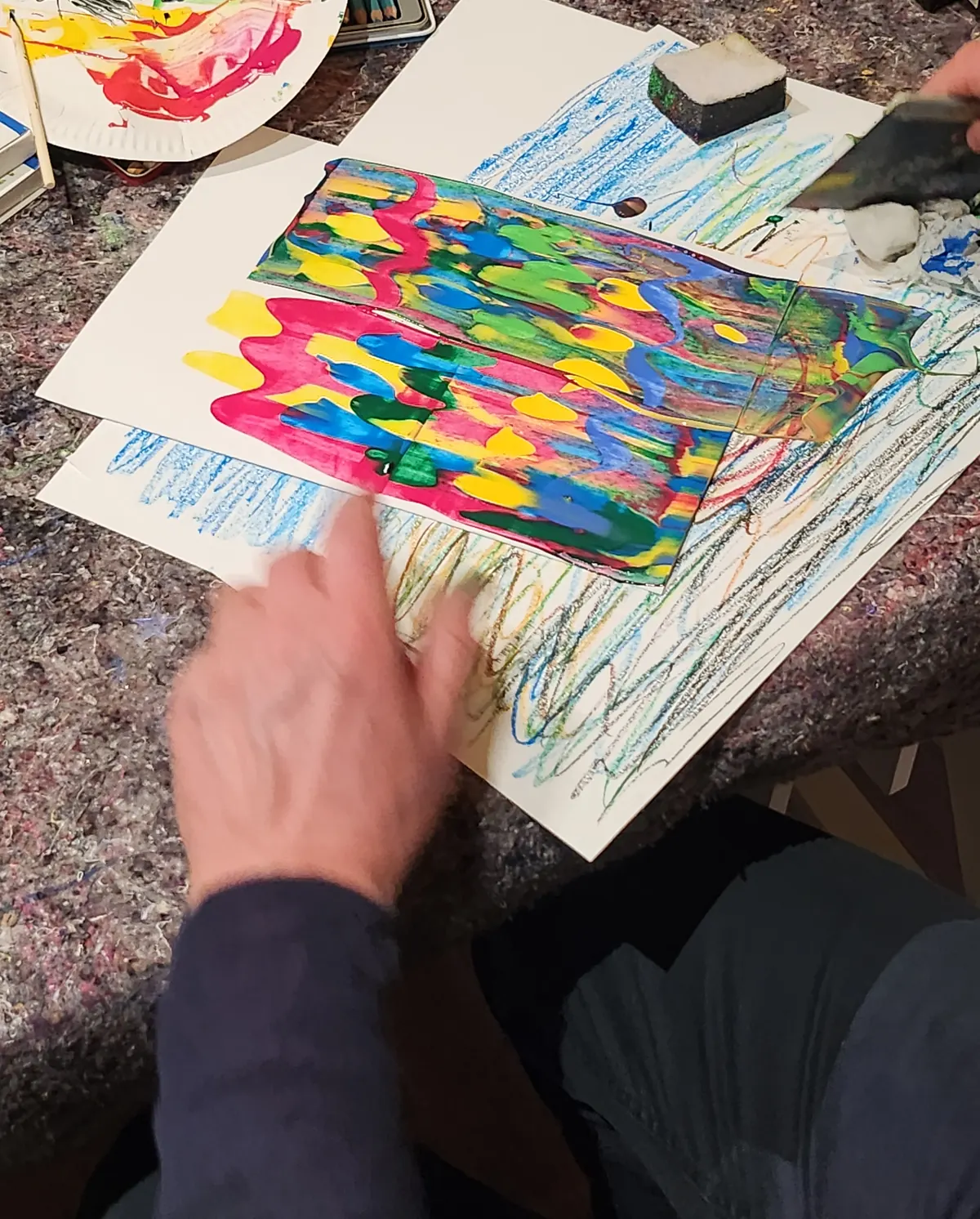 Händer arbetar med ett färgglatt tryck och pastellkritor under en kreativ kompiskväll.